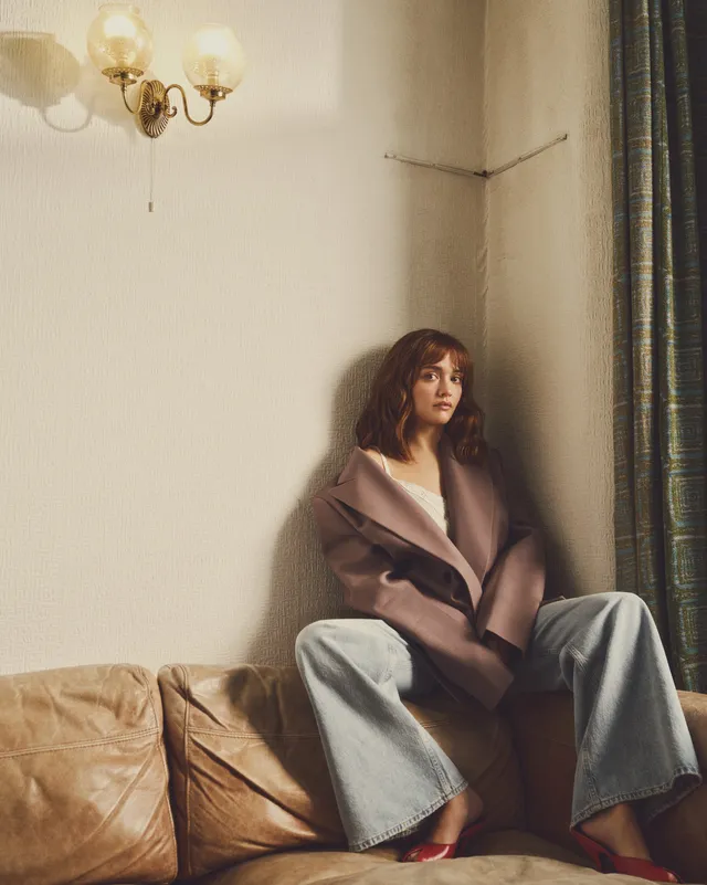 Olivia Cooke sitting on a sofa in loose jeans.