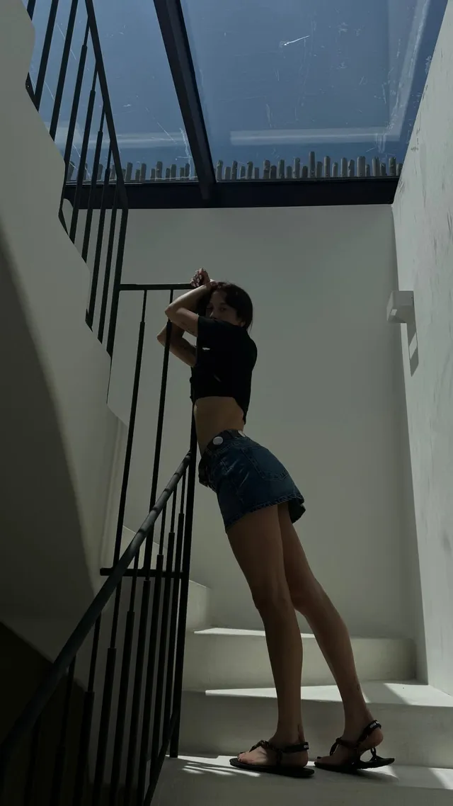 María Pedraza posing on stairs in a dark top, denim shorts and flat sandals.