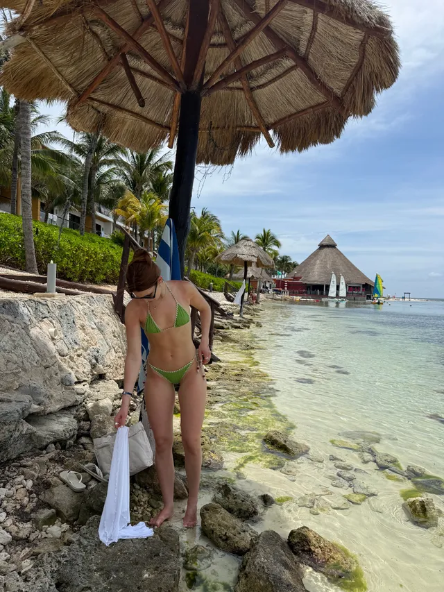 Sophie Nélisse standing barefoot on rocks beside clear sea water.