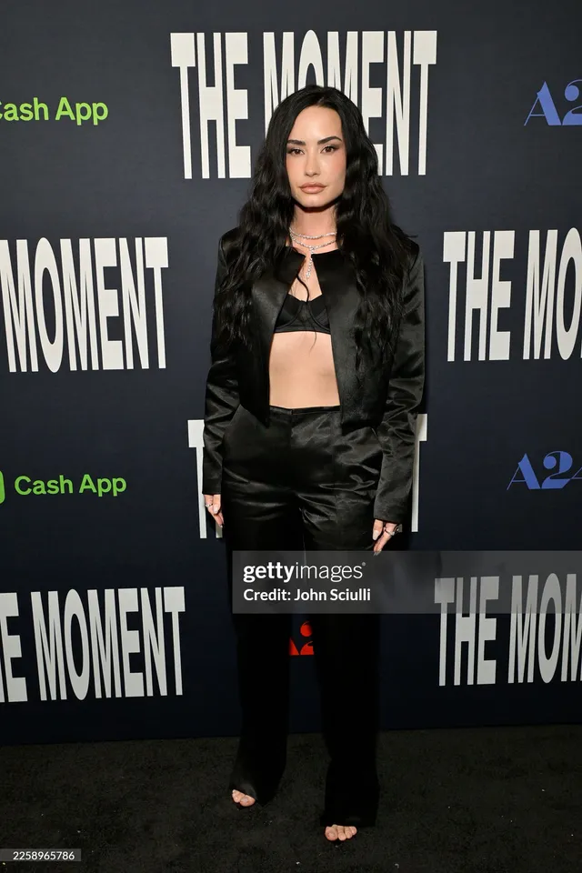 Demi Lovato posing at an event in a black outfit and open-toe footwear.