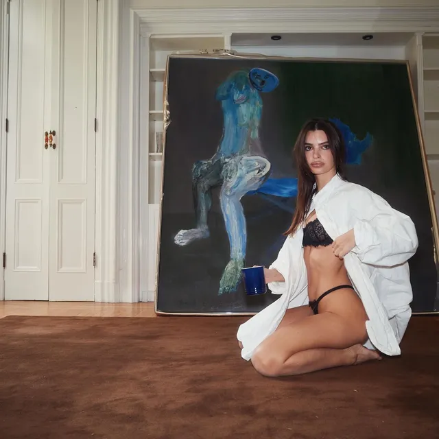 Emily Ratajkowski kneeling barefoot on a rug beside a painting.