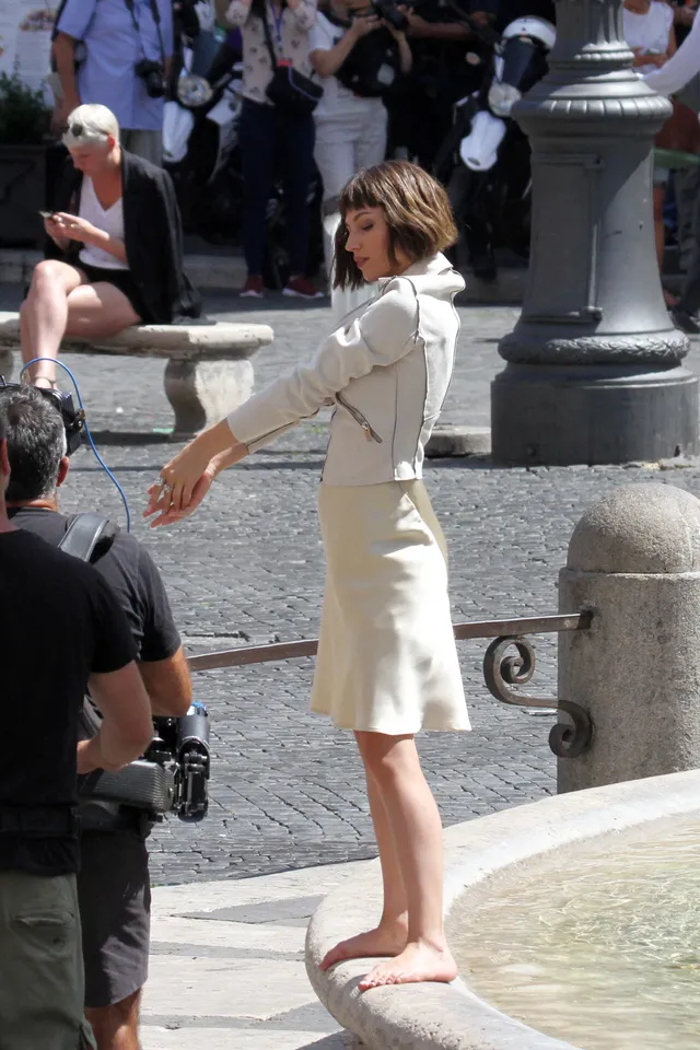 Úrsula Corberó standing barefoot on a stone fountain edge in a pale dress.