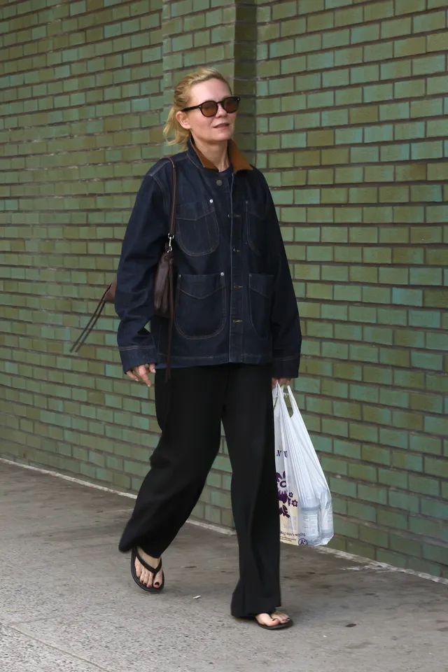 Kirsten Dunst walking outside in a dark jacket and sandals.
