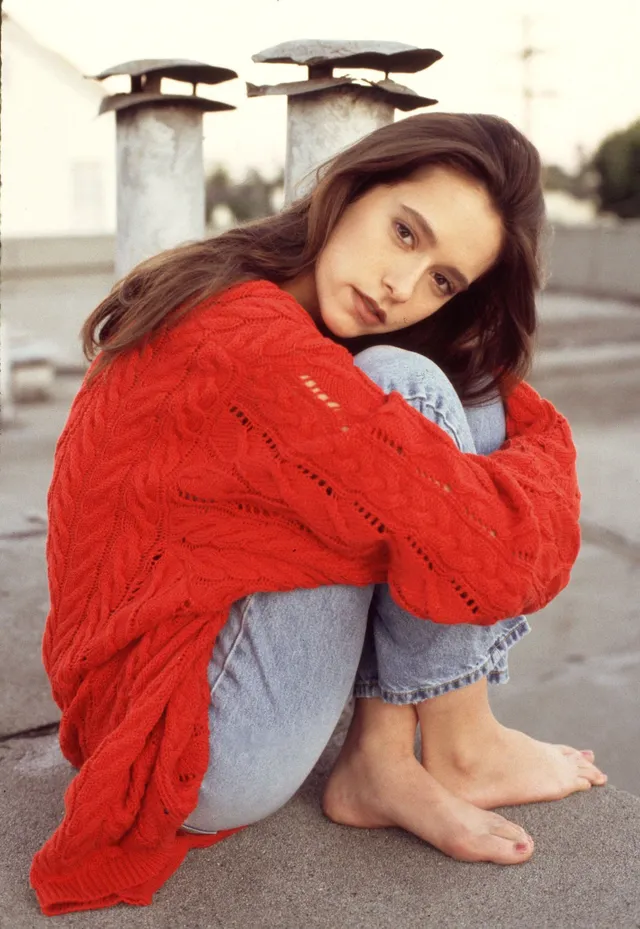 Jennifer Love Hewitt sitting barefoot outdoors in a red sweater and jeans.