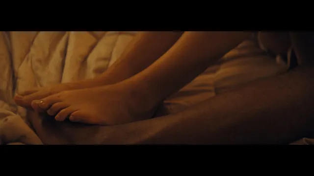 Gemma Chan resting bare feet on bedding in a screen scene.