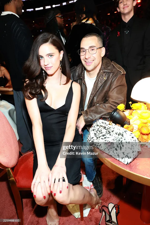 Margaret Qualley seated at an event with bare soles visible.
