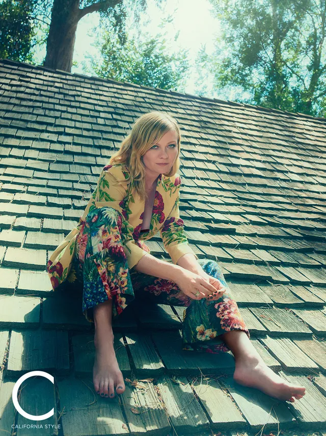 Kirsten Dunst sitting barefoot on a roof in a floral outfit.