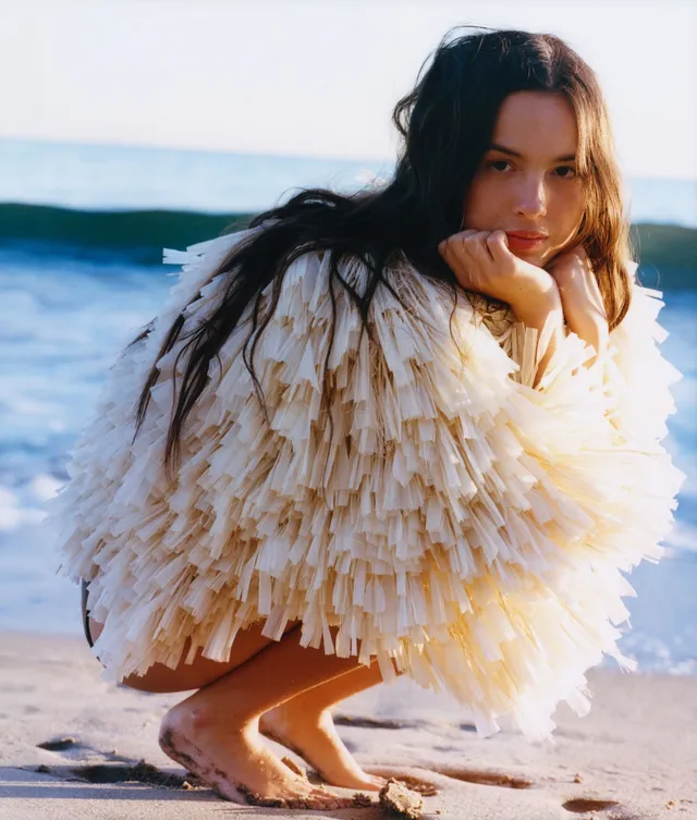 Olivia Rodrigo crouching beside water in a feathered outfit.