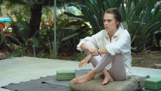 Cara Delevingne sitting outdoors in a white shirt beside yoga equipment.