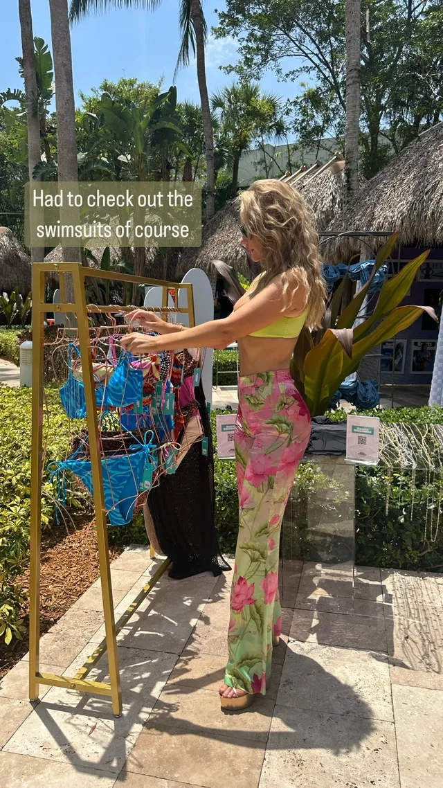 Paige Spiranac standing in heeled sandals and floral trousers beside tropical plants.