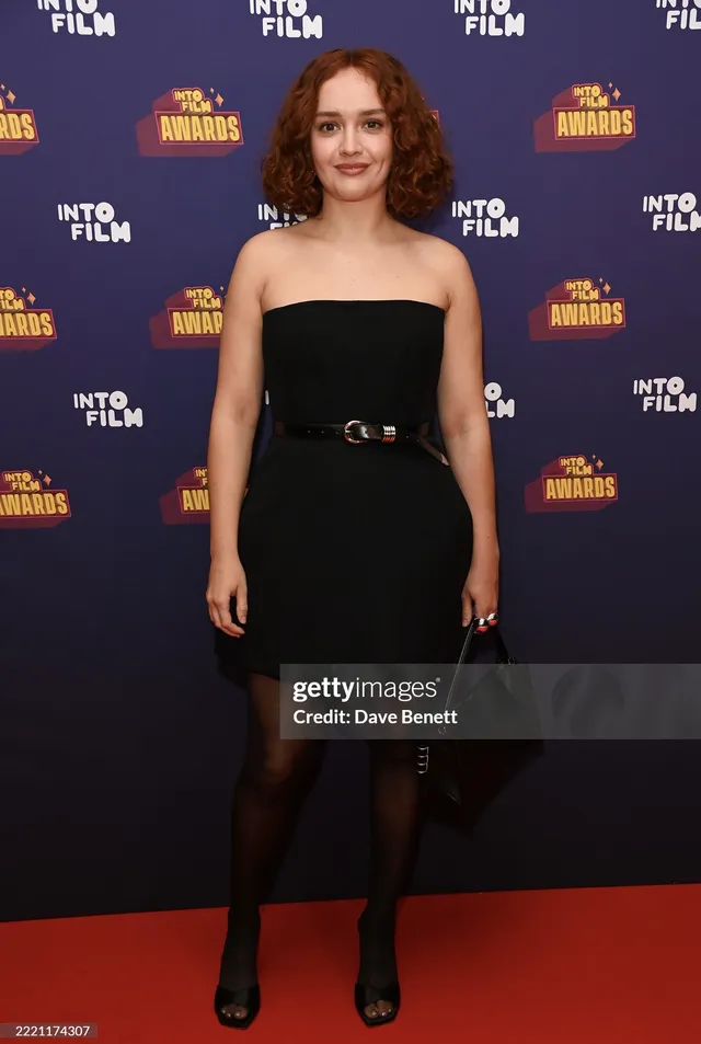 Olivia Cooke posing in a short black dress and boots.