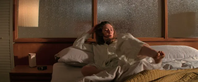 Elizabeth Olsen sitting barefoot on a bed in a loose outfit.