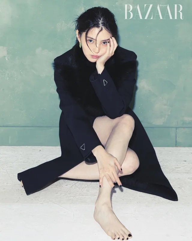 Han So-hee sitting barefoot in a dark coat.