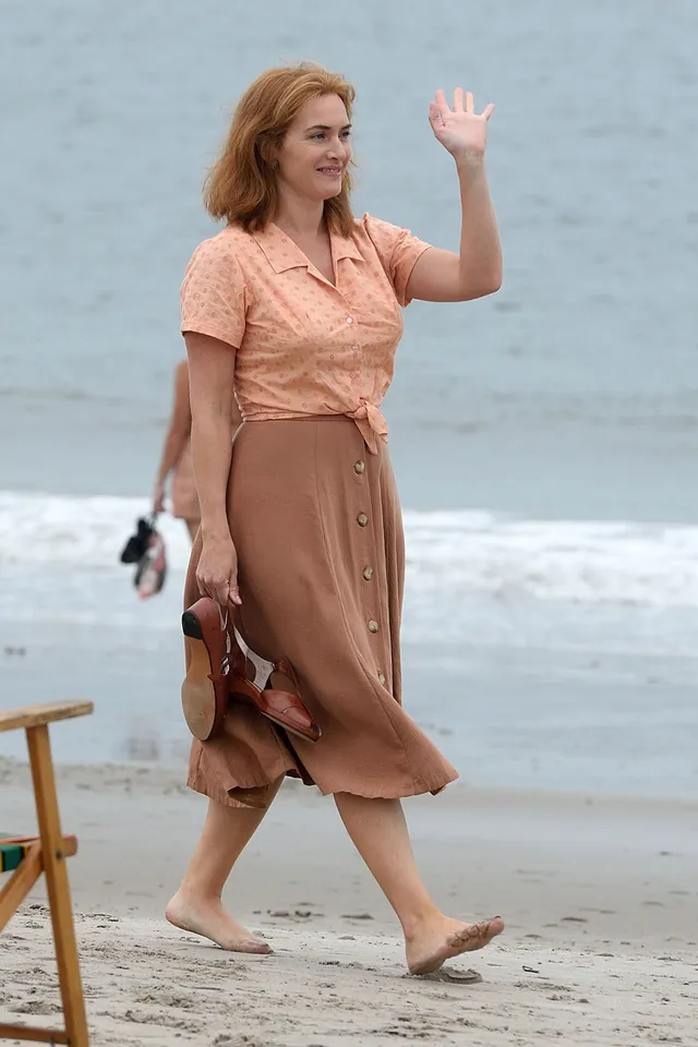 Kate Winslet walking barefoot on a beach in a peach dress.