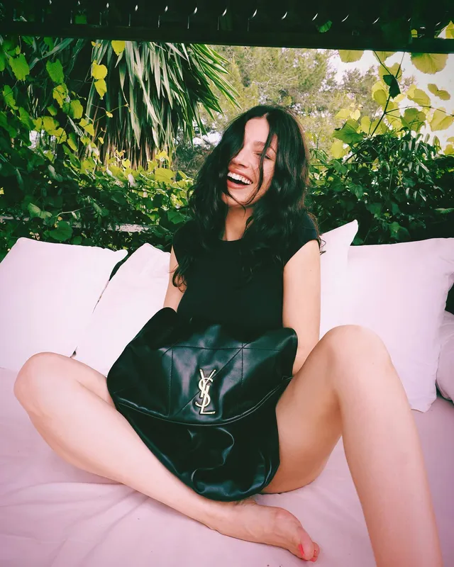 Úrsula Corberó sitting barefoot on a pink sofa in a black outfit.