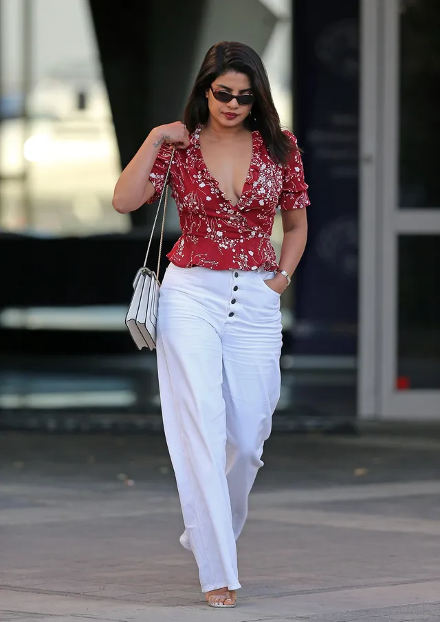 Priyanka Chopra Jonas walking in pale heels, white trousers and a patterned red top.