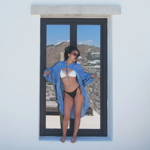 Rachel Zegler standing barefoot in a doorway in a swimsuit and blue shirt.