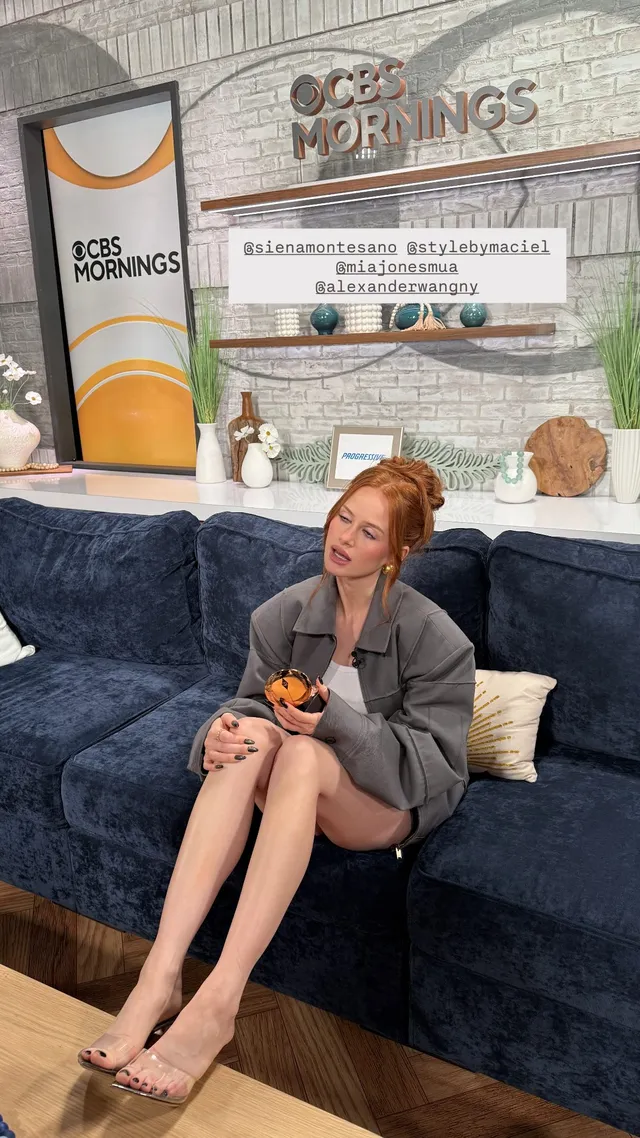 Madelaine Petsch sitting on a blue sofa in a gray outfit and heels.