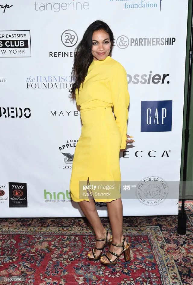 Rosario Dawson posing indoors in a yellow dress.