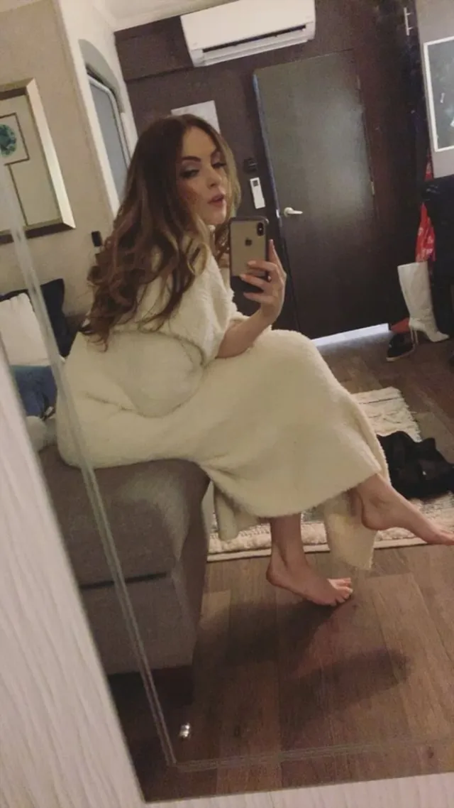 Elizabeth Gillies taking a seated mirror portrait in a pale dress.