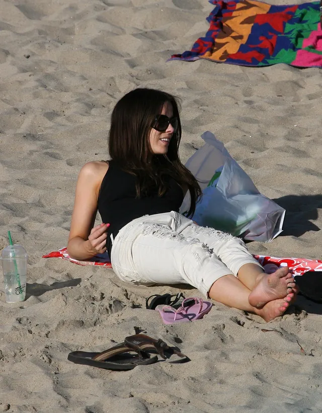 Kate Beckinsale sitting on a beach with bare soles facing the camera.