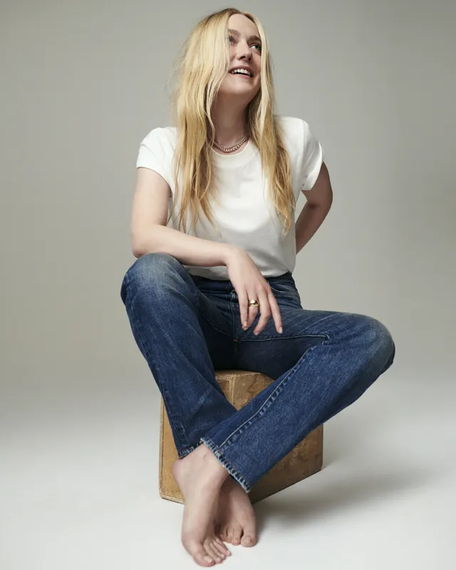 Dakota Fanning sitting barefoot on a stool in jeans and a white shirt.