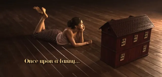 Deepika Padukone lying on a wooden floor beside a model house with her bare feet raised.
