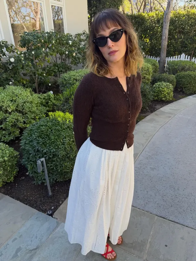 Leighton Meester standing outdoors in a brown cardigan and white skirt.
