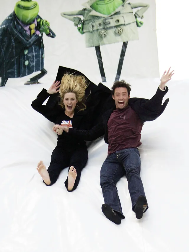 Kate Winslet reclining barefoot beside a companion on a white background.