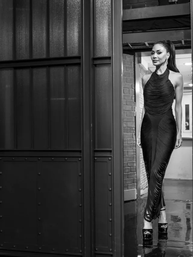Nicole Scherzinger standing indoors in a black gown and platform heels.