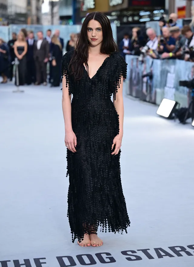 Margaret Qualley posing on a pale carpet in a black dress and heels.