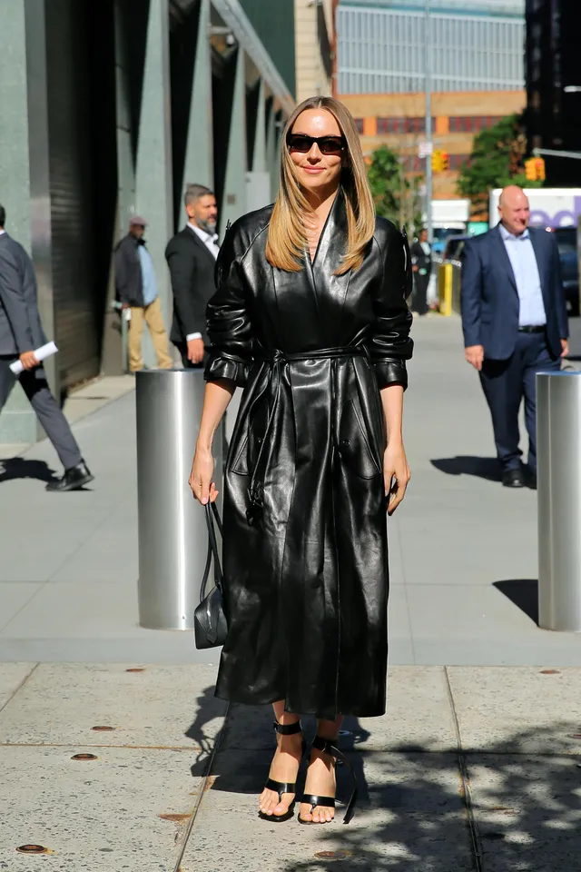 Jodie Comer standing on a sidewalk in a long black coat.
