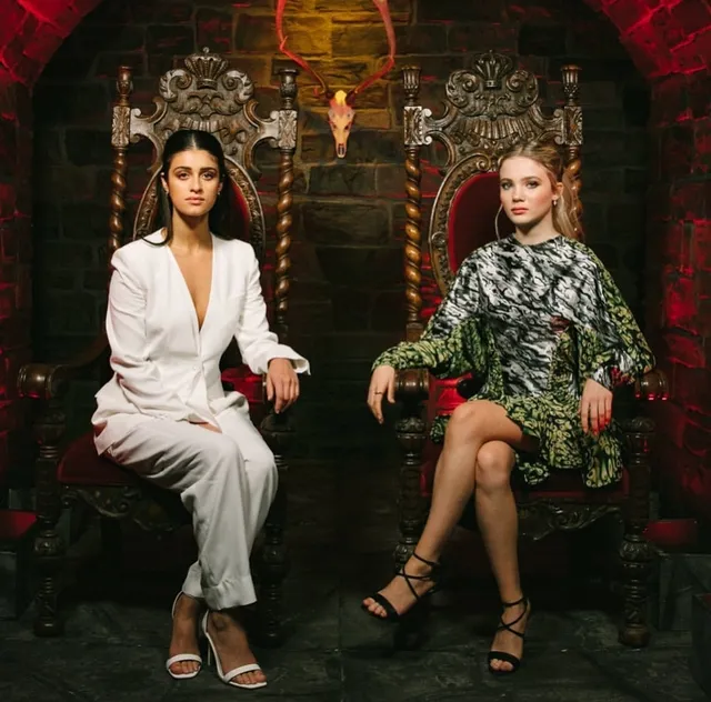 Anya Chalotra sitting beside another woman in a white suit and pale heels.