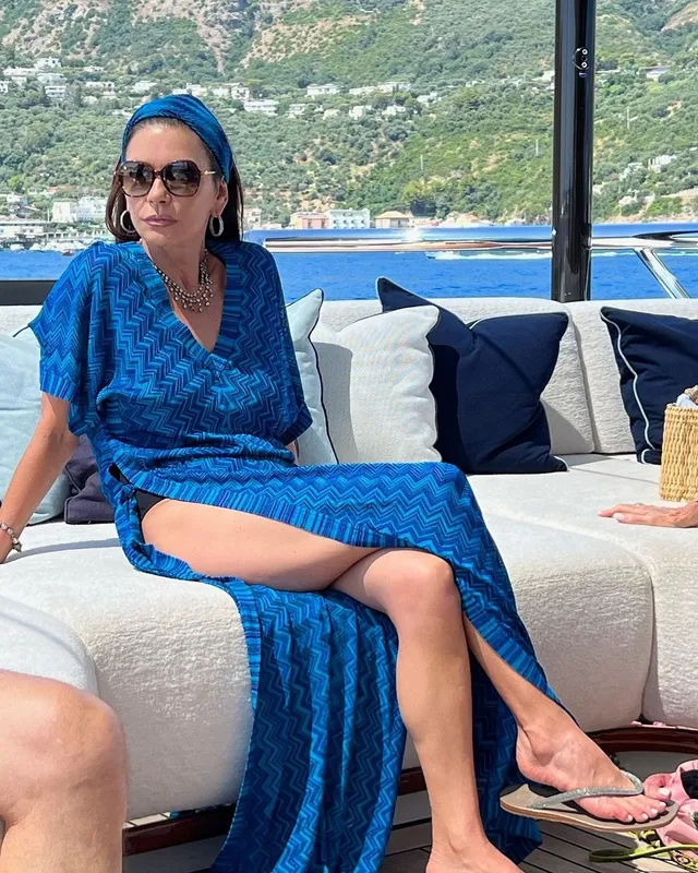 Catherine Zeta-Jones seated outdoors in a blue outfit with flat sandals.