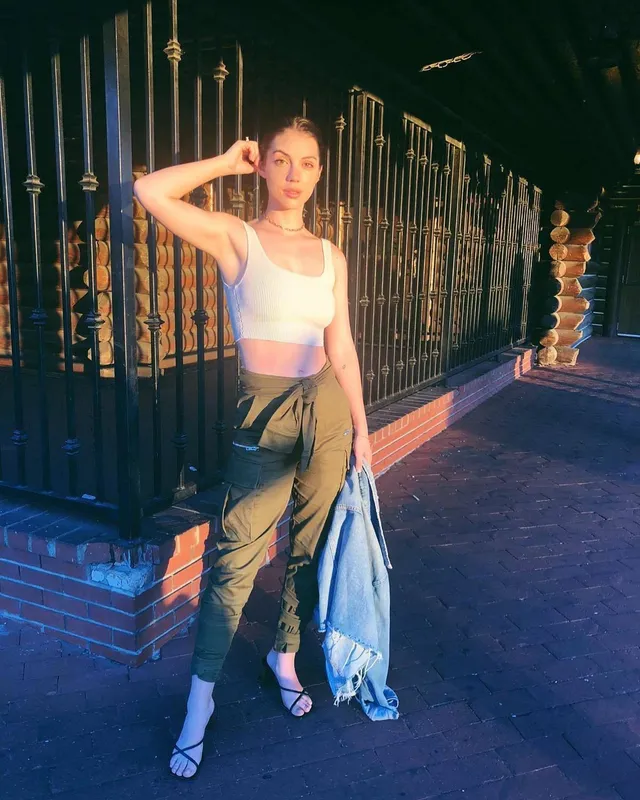 Adelaide Kane standing beside a gate in cargo trousers and heels.