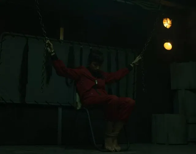 Úrsula Corberó sitting barefoot in a red jumpsuit in a dimly lit scene.