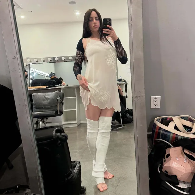 Billie Eilish a cream outfit and white leg warmers in a mirror portrait.