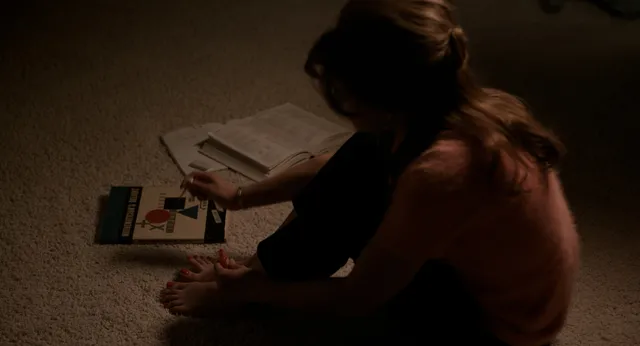 Cailee Spaeny sitting barefoot on a carpet beside open books.