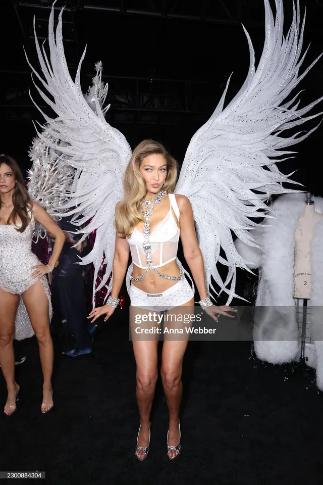 Gigi Hadid standing backstage in a white winged outfit and heels.