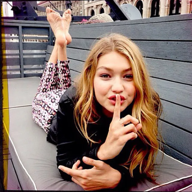 Gigi Hadid reclining with bare soles raised on an outdoor cushion.