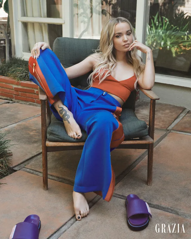 Dove Cameron sitting barefoot in blue trousers with slippers nearby.