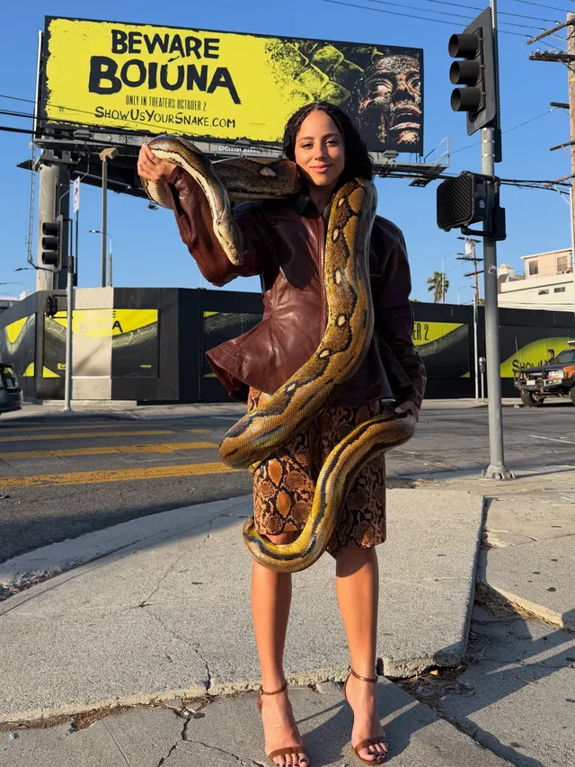 Kiana Madeira posing outdoors with a snake in a patterned dress and brown heels.
