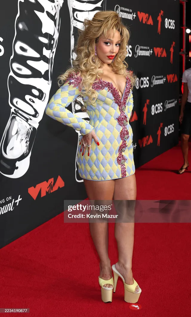 Doja Cat posing at an event in a patterned dress and gold platform heels.
