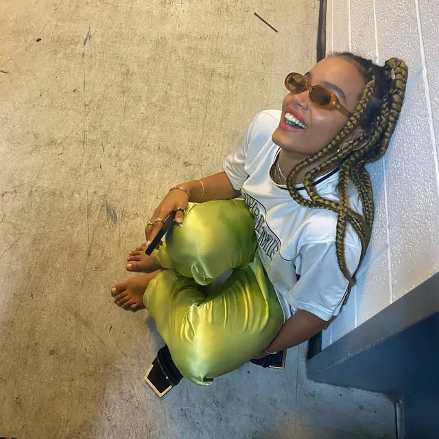 Whitney Peak sitting indoors in a white T-shirt, metallic green skirt and heels.