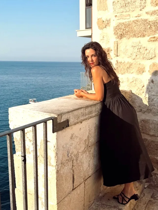 Danielle Campbell leaning against a seaside balcony in a black dress.