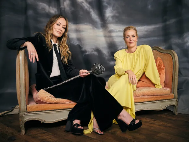 Olivia Wilde sitting with one bare foot on a sofa beside another person.