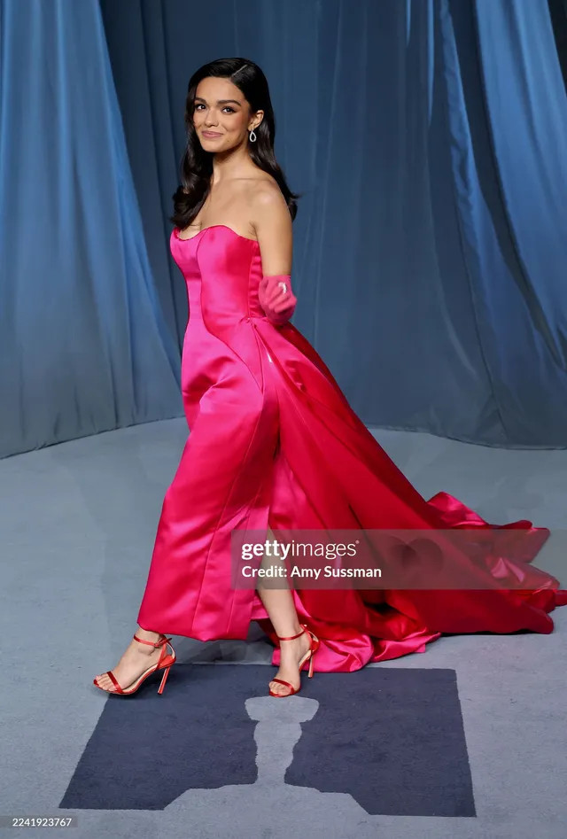 Rachel Zegler turning on a carpet in a pink gown and red heels.