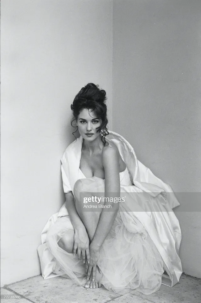 Monica Bellucci sitting barefoot in pale fabric for a black and white portrait.