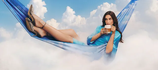 Dua Lipa reclining in a blue hammock wearing heeled sandals.