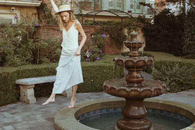Phoebe Dynevor posing beside a garden fountain in a pale dress.